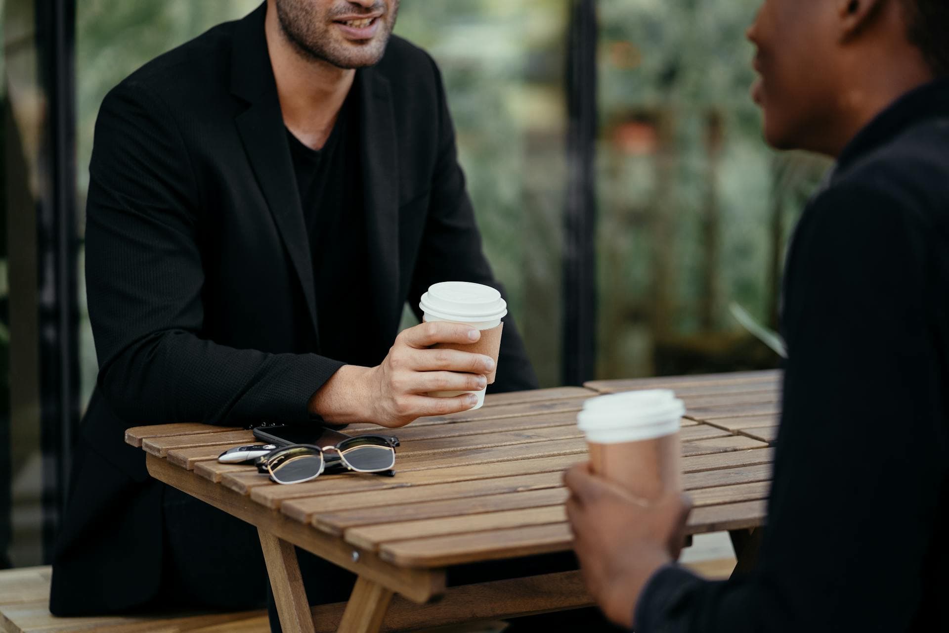 Two men talking over takeaway coffee at a wooden table, faces out of frame.