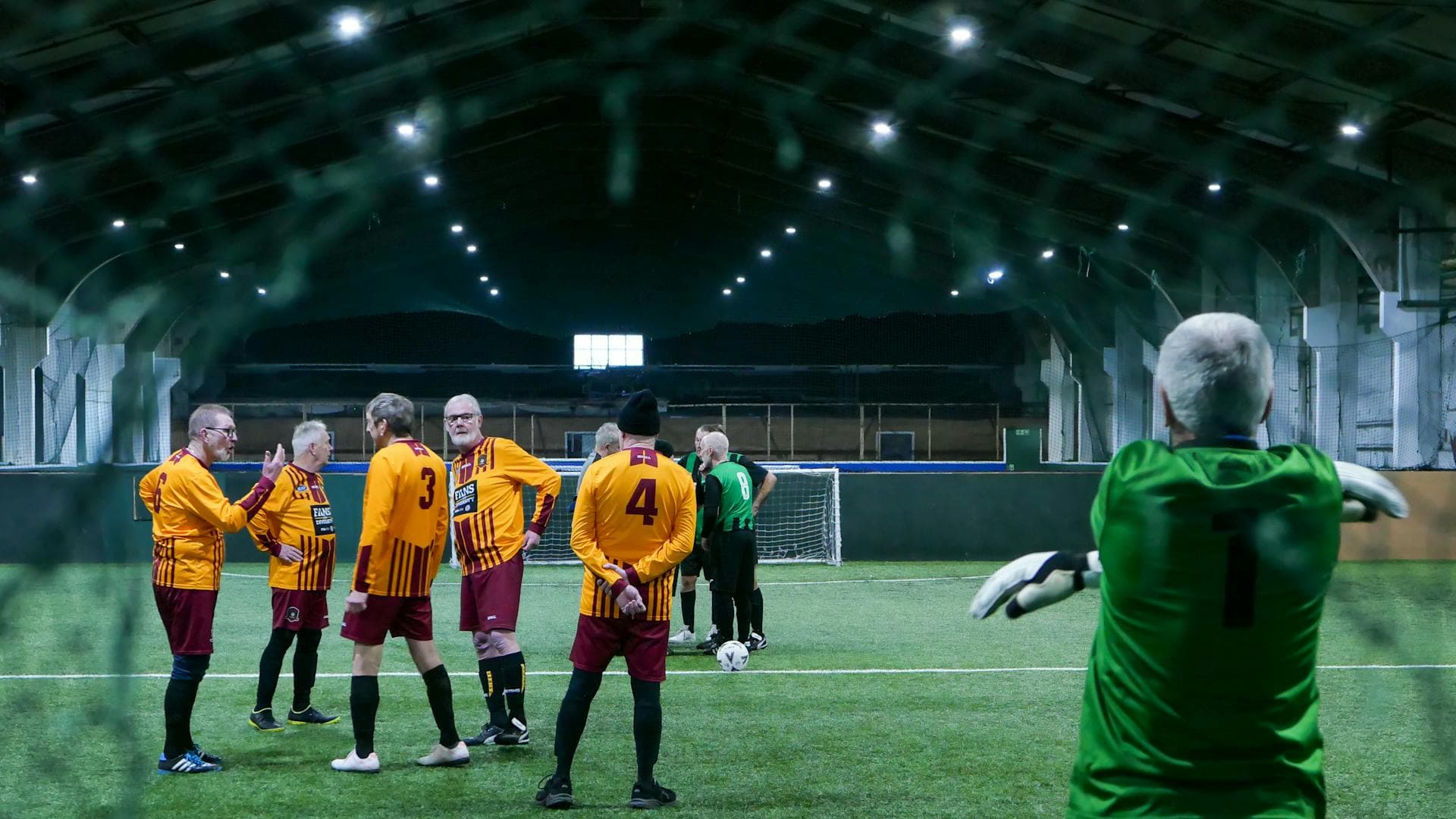 Older men playing indoor walking football at a UK sports centre.