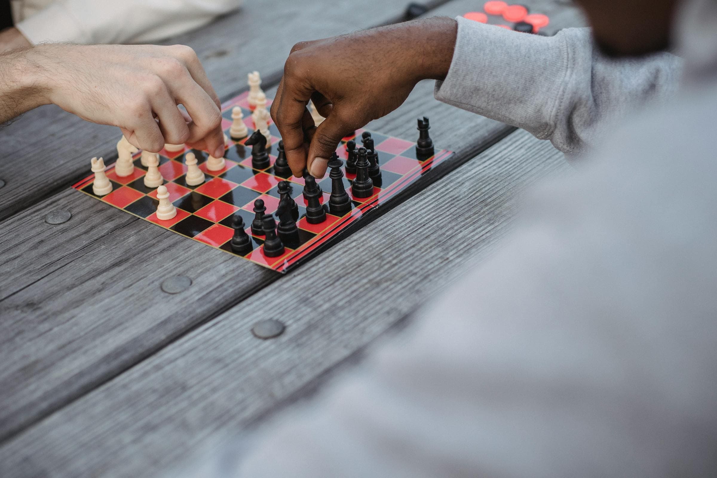 Close-up from above of two men's hands, one Black and one white, moving pieces on a chessboard across an outdoor wooden table.